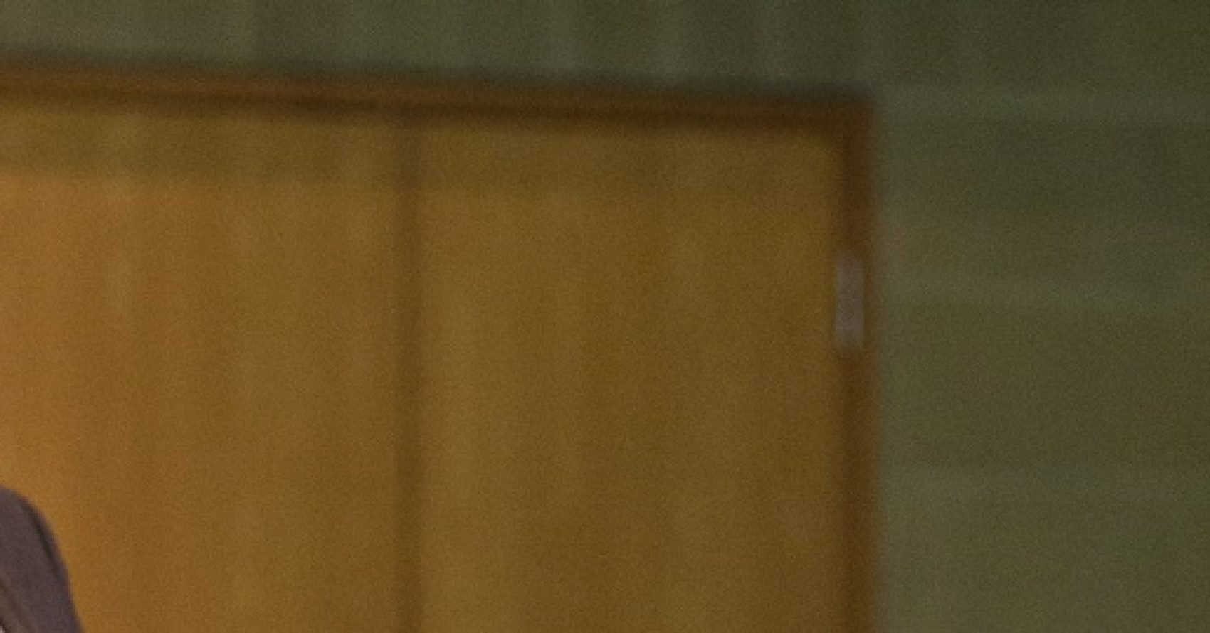 A man wearing a suit stands indoors speaking, with a wooden door and a blurred background behind him.