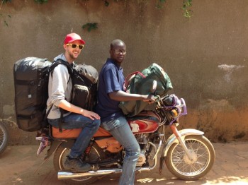 Two men on a motorbike in Africa - one a UK volunteer in sunglasses and a baseball cap, the other a local in reading glasses.