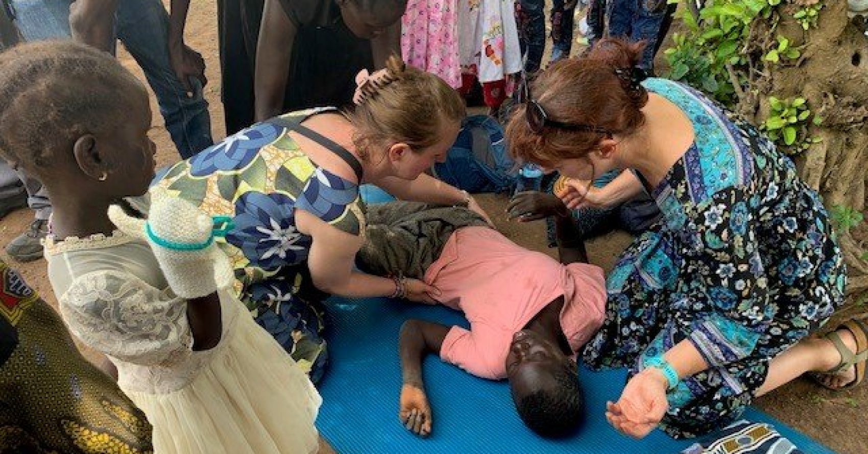 Two female UK therapists help a child who is lying on a mat outside while several people observe in the background.