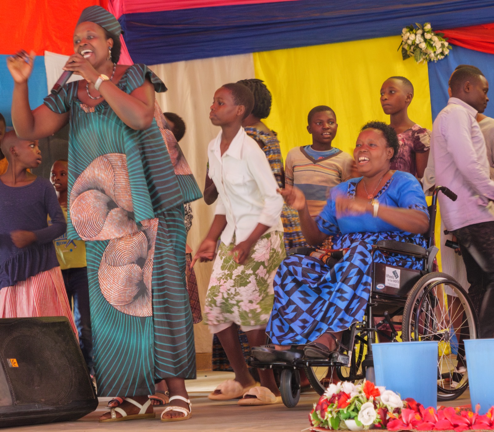 People, including a woman in a wheelchair, sing and dance together on a vibrant, colorful stage.