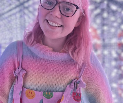 A person, Triona Brading, with pink hair and glasses smiles, dressed in colourful overalls with smiley faces and a rainbow-striped sweater.