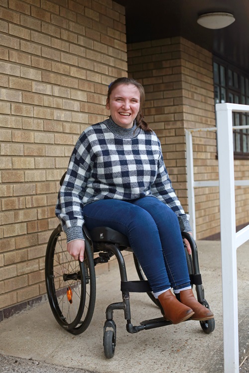 A woman on her ABC Journey smiles while sitting in a wheelchair outdoors next to a brick wall and white railing.