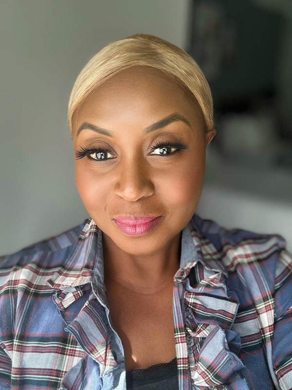 A blonde-haired woman in a plaid shirt smiles gently at the camera, illuminated by natural lighting.