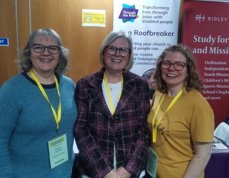 Three women with name badges smile and stand together in front of informational banners at an indoor event.