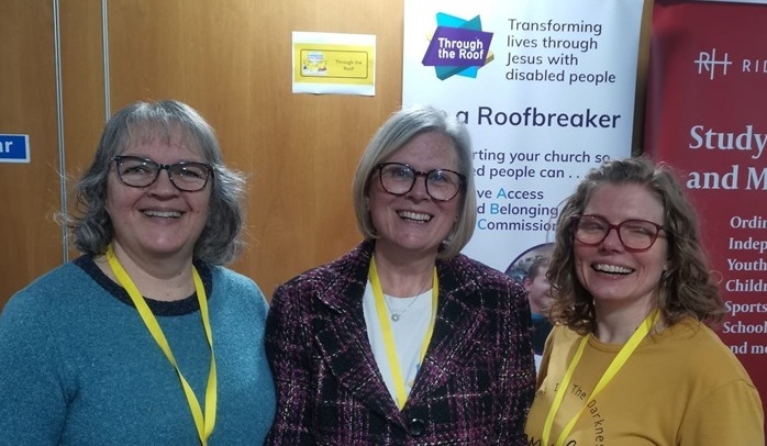 Three women wearing glasses and yellow lanyards stand together in front of a Roofbreaker banner at an indoor event, all smiling.