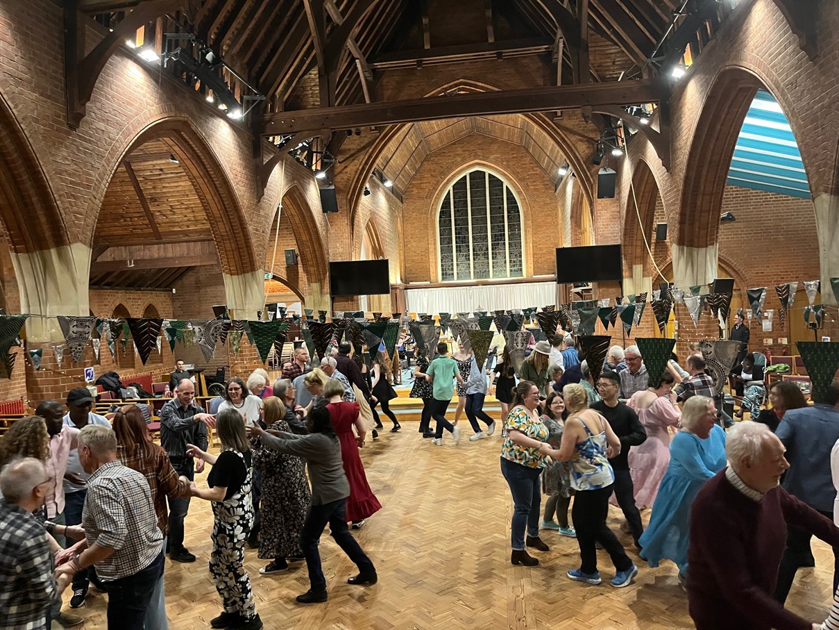 A large group of people of all ages dance in a church sanctuary in Ashtead linked to St George's and St Giles. Bunting adorns and ornate decorations, with high ceilings, and arched windows during a fundraising barn dance event.