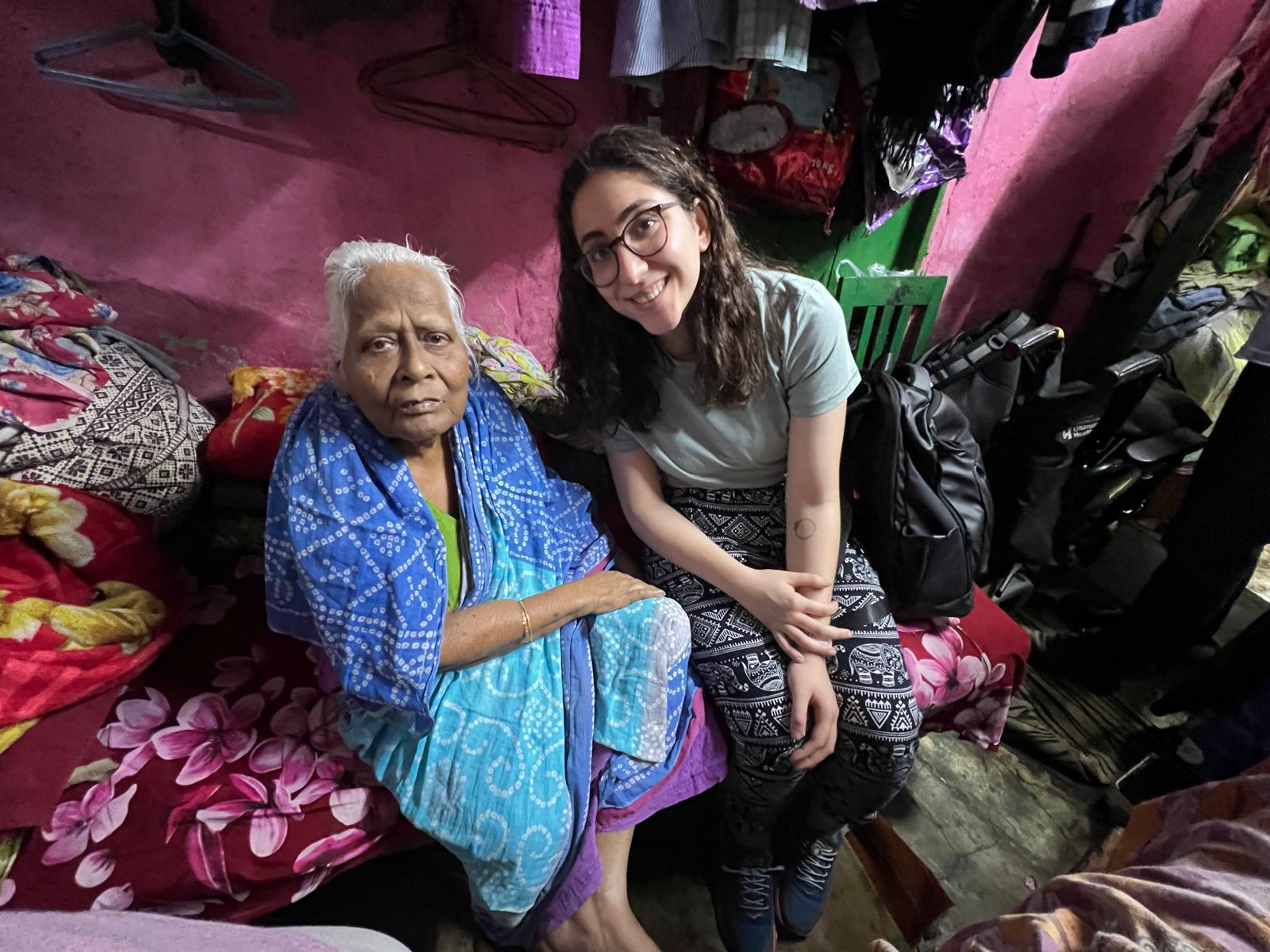 A young UK woman and an elderly Indian woman sit side by side on a bed in a colorful room, both smiling at the camera.